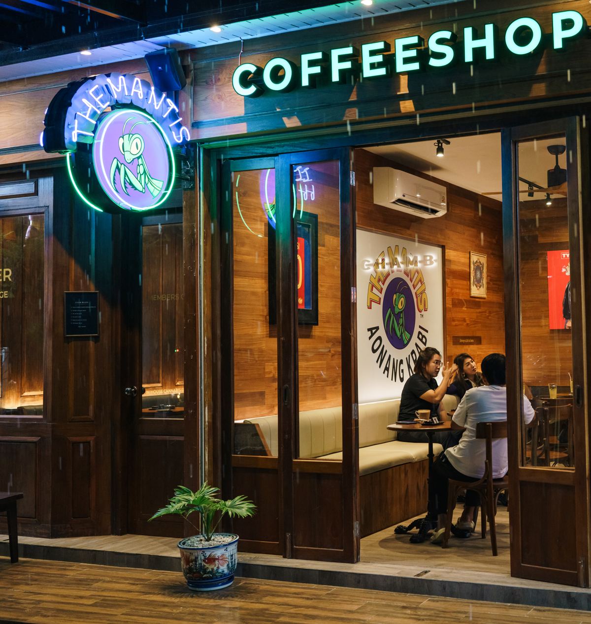 Front of Coffeeshop The Mantis at night: neon mantis logo, glass doors and guests chatting on the cream banquette inside